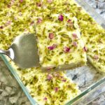 A close-up shot of Ayshe Elsaraya, a Middle Eastern dessert with layers of golden toasted bread soaked in sweet syrup, topped with a creamy ashta layer, crushed pistachios, and decorative rose petals.