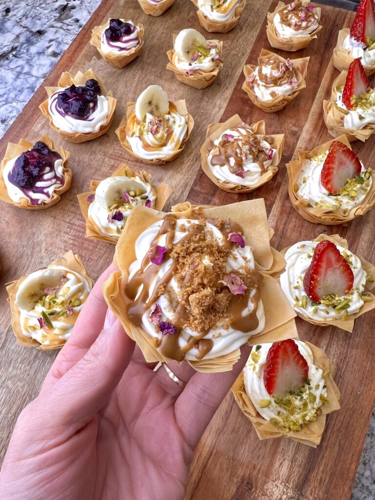 Baklava cheesecake bites drizzled with smooth caramel sauce and topped with a swirl of whipped cream, adding a rich, sweet touch.
