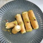 Golden brown Ashta Baklava Rolls made with crispy phyllo dough, filled with creamy Arabic ashta, drizzled with floral simple syrup, and garnished with crushed pistachios, arranged elegantly on a serving plate.