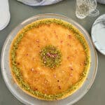 Golden and crispy baked othmaliye topped with crushed pistachios, drizzled with simple syrup, and showcasing layers of shredded kataifi dough, creamy ashta filling, and melted mozzarella cheese