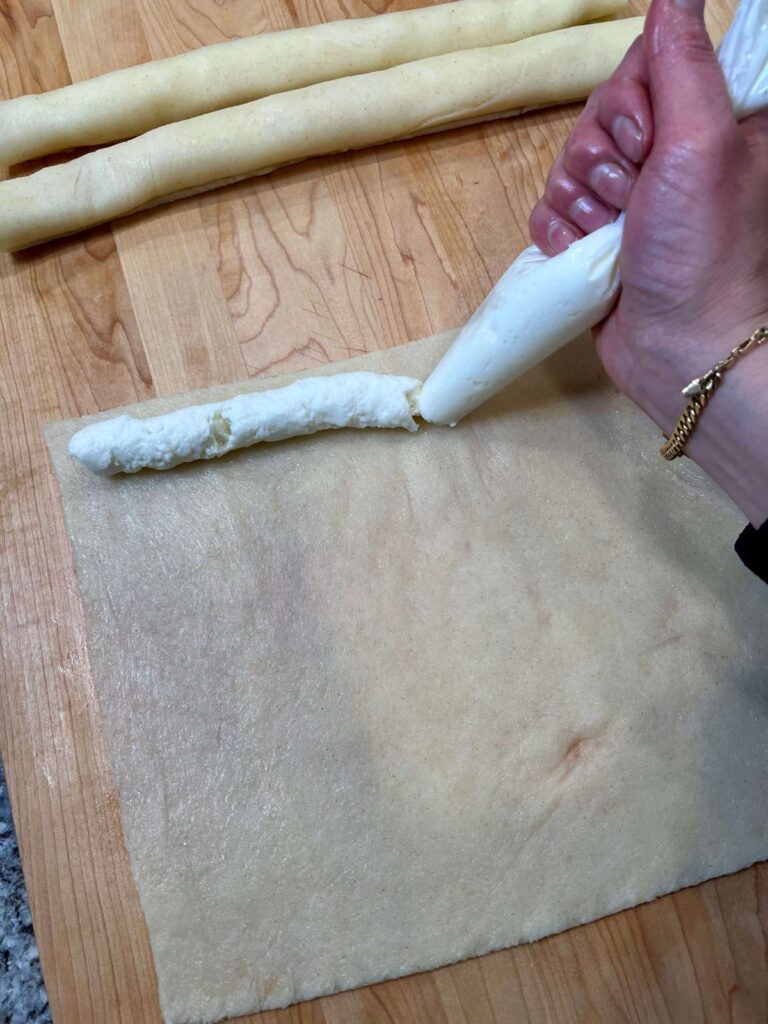A piping bag filled with creamy ashta, squeezing a line of filling onto the cheese dough.