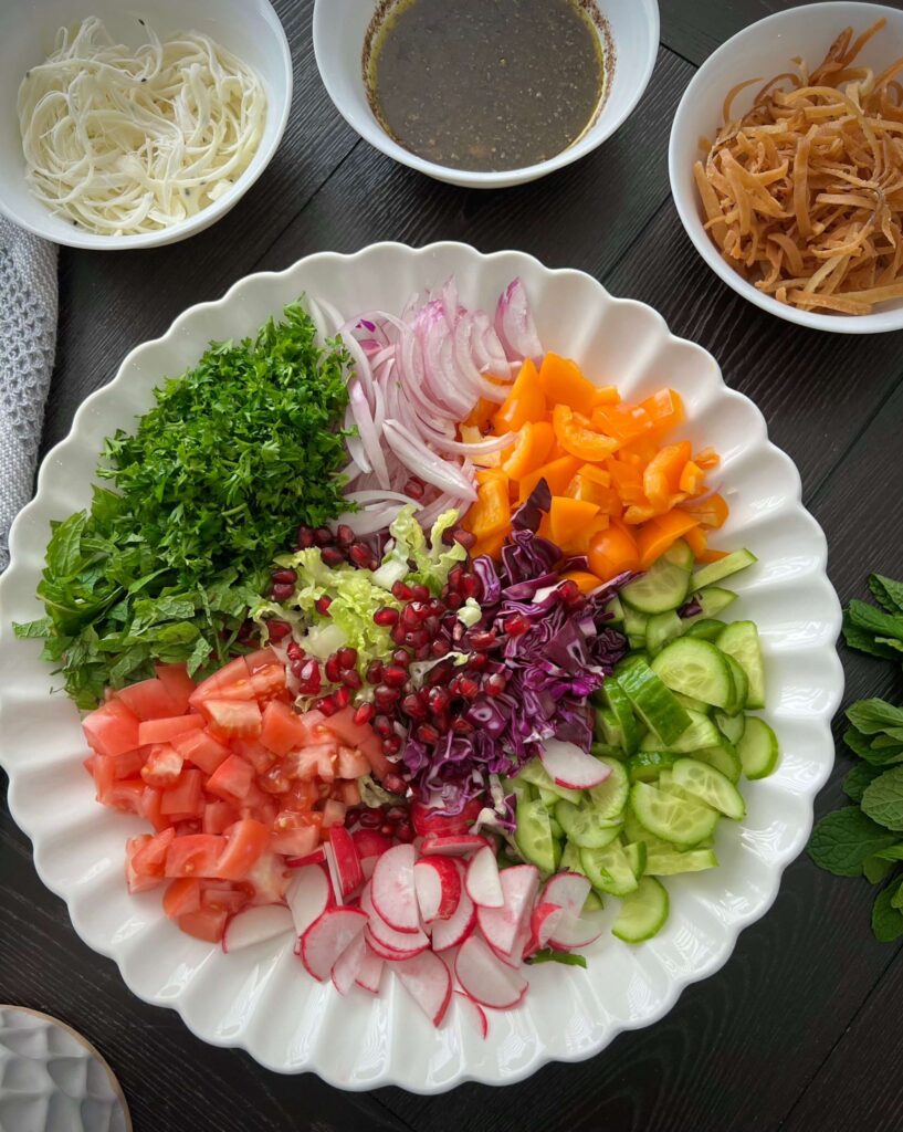 Chopped vegetables including romaine lettuce, cucumbers, tomatoes, radishes, sweet peppers, red onion, and fresh herbs like parsley and mint, prepared for a salad.