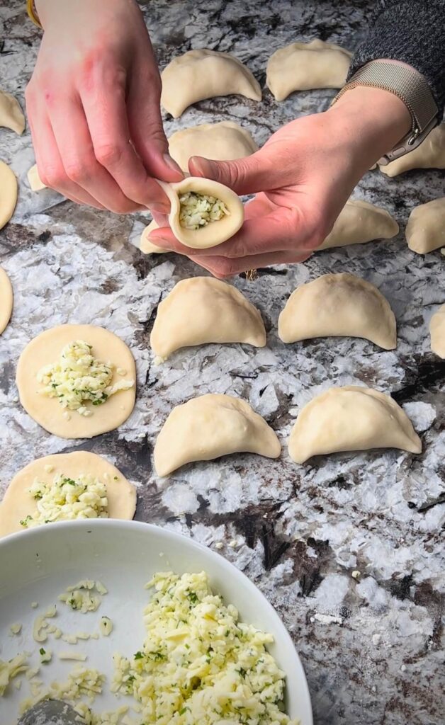 A hand placing a spoonful of cheese filling onto a rolled-out dough circle, preparing to fold and seal the cheese sambousek.