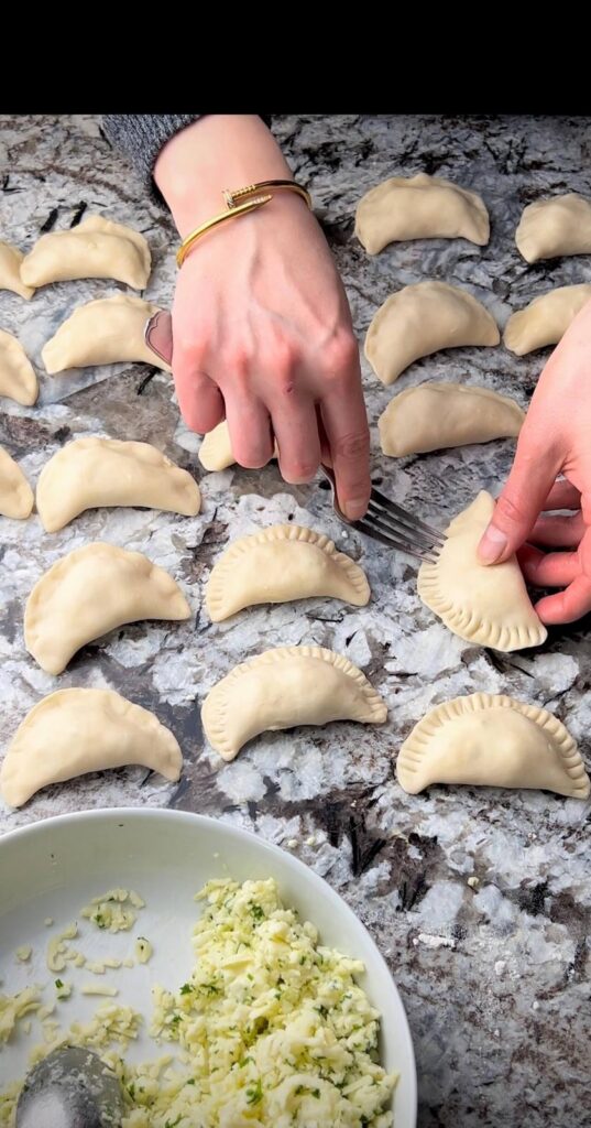 A hand placing a spoonful of cheese filling onto a rolled-out dough circle, preparing to fold and seal the cheese sambousek.