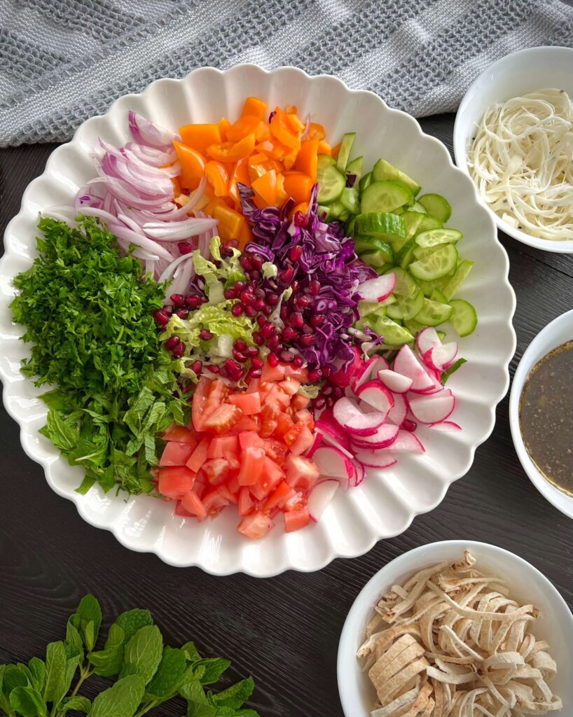 Chopped vegetables including romaine lettuce, cucumbers, tomatoes, radishes, sweet peppers, red onion, and fresh herbs like parsley and mint, prepared for a salad.