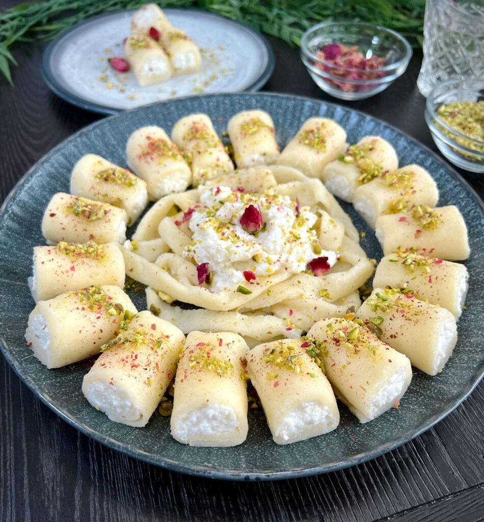 A plate of Halawet el Jibn rolls, showcasing soft, golden cheese dough filled with creamy ashta, garnished with crushed pistachios and rose petals, and drizzled with glossy simple syrup.