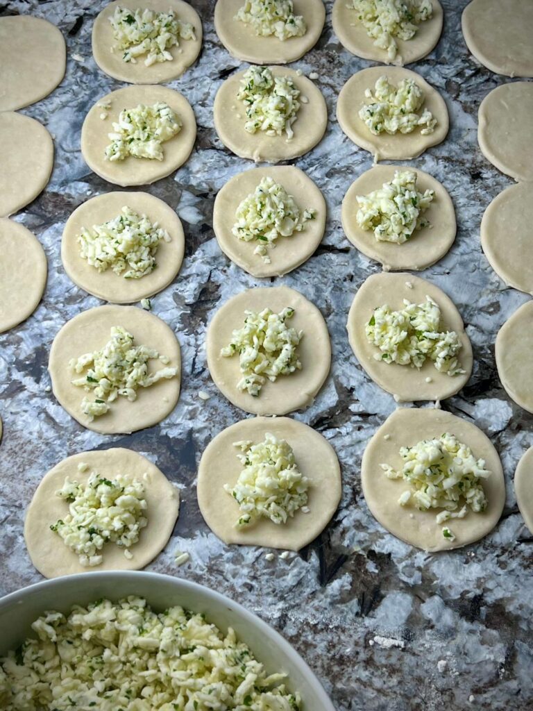 A hand placing a spoonful of cheese filling onto a rolled-out dough circle, preparing to fold and seal the cheese sambousek.