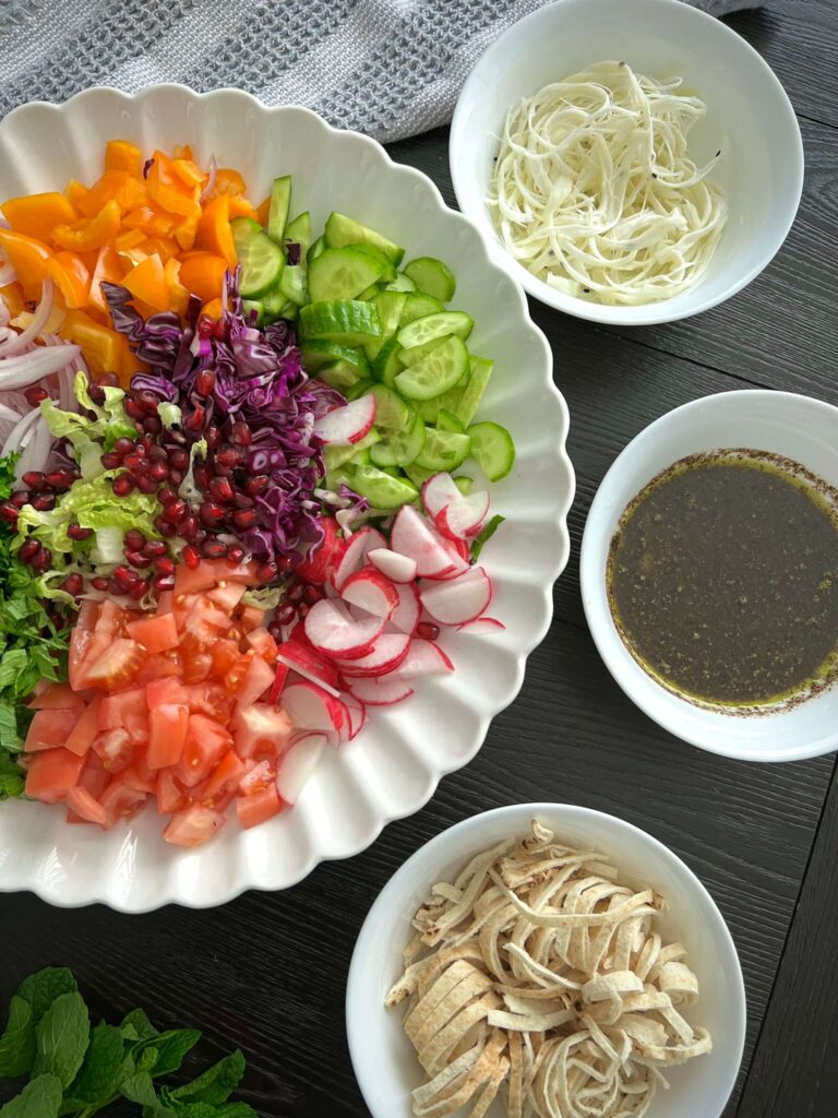 Chopped vegetables including romaine lettuce, cucumbers, tomatoes, radishes, sweet peppers, red onion, and fresh herbs like parsley and mint, prepared for a salad.