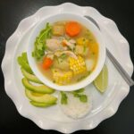 A bowl of Colombian Chicken Sancocho with tender chicken, yuca, plantains, corn on the cob, and potatoes in a rich golden broth, garnished with fresh cilantro and served with a side of rice and avocado.