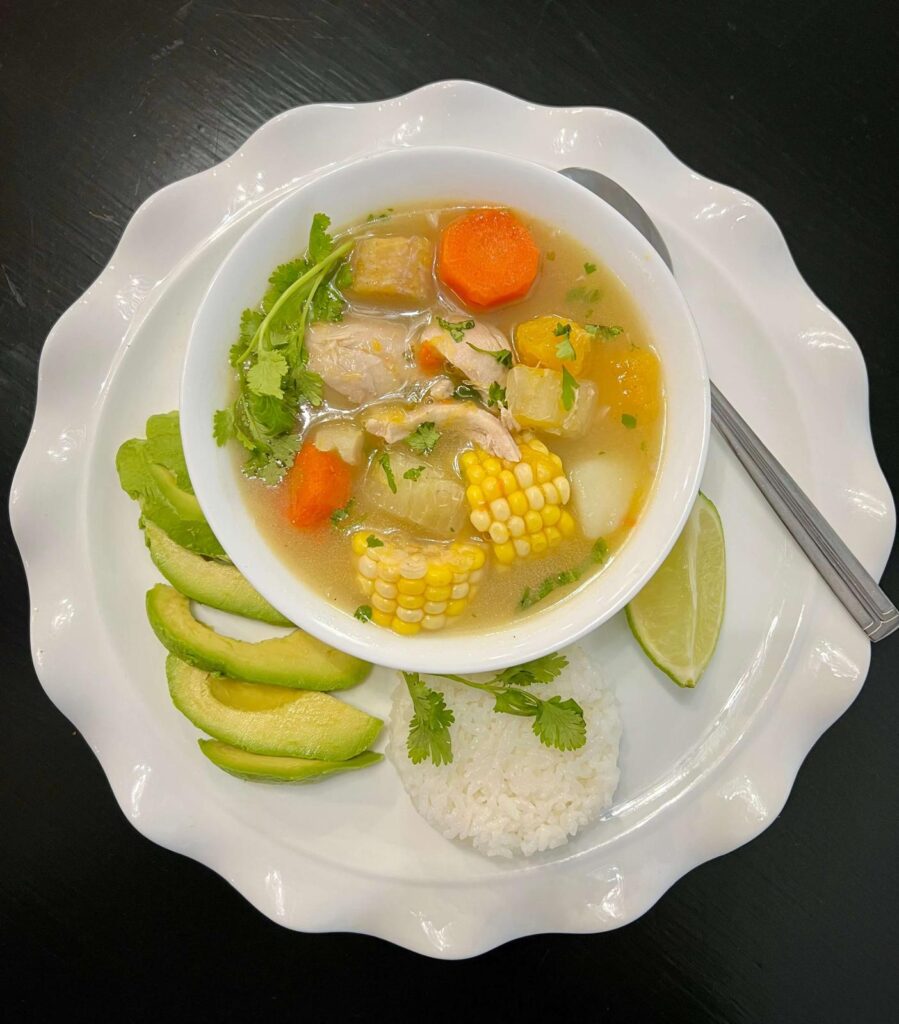 A bowl of Colombian Chicken Sancocho with tender chicken, yuca, plantains, corn on the cob, and potatoes in a rich golden broth, garnished with fresh cilantro and served with a side of rice and avocado.